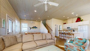 A spacious living area with a beige sofa, a blue armchair with a nautical theme, a wooden coffee table, and a kitchen area with white cabinets and a microwave. - GV634, Shared Pool, Pet Friendly, Charcoal Grill, Close to Beach (Port Aransas)