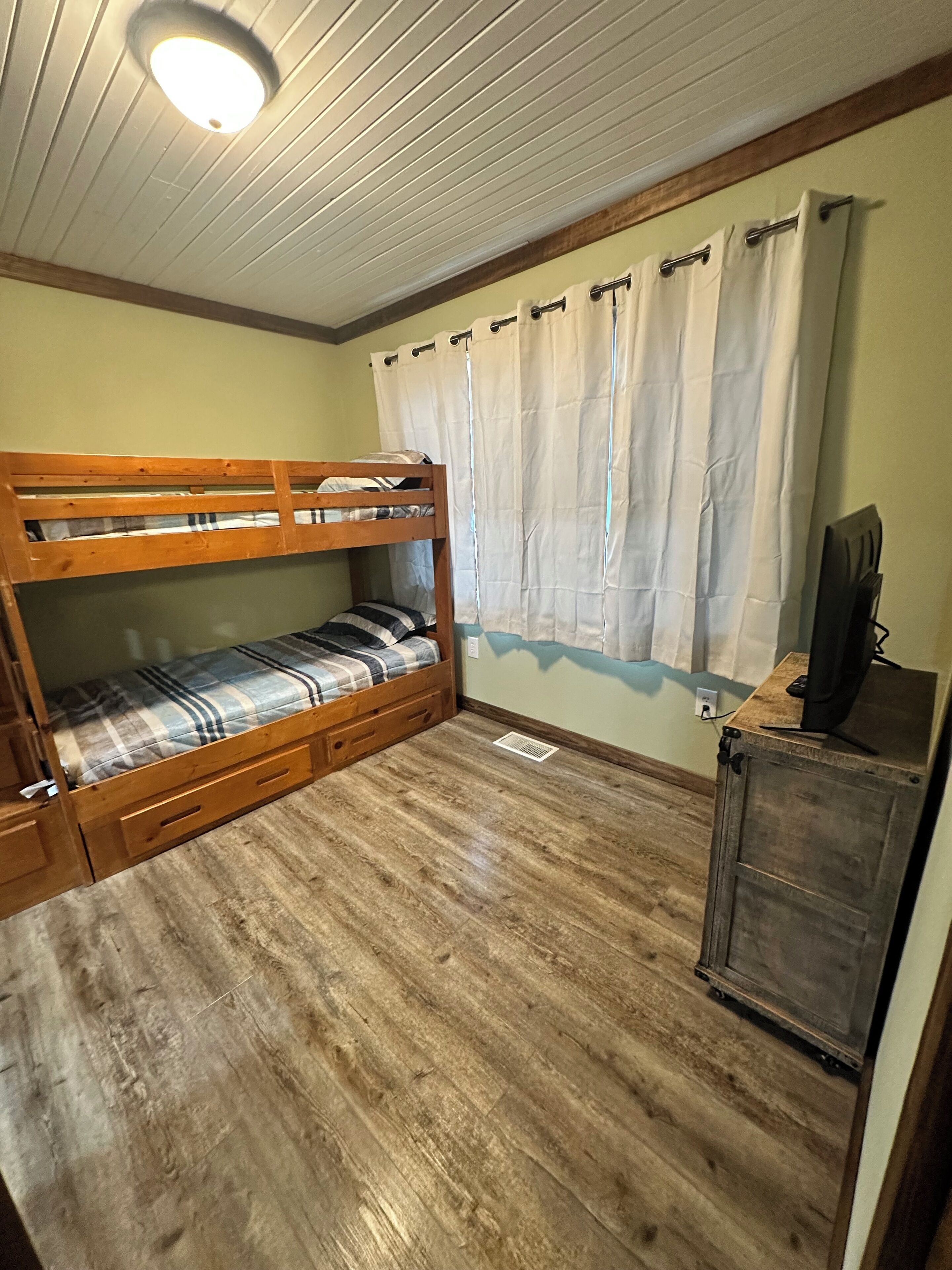 Une chambre avec un lit superposé, un plancher en bois, un luminaire au plafond et un rideau.