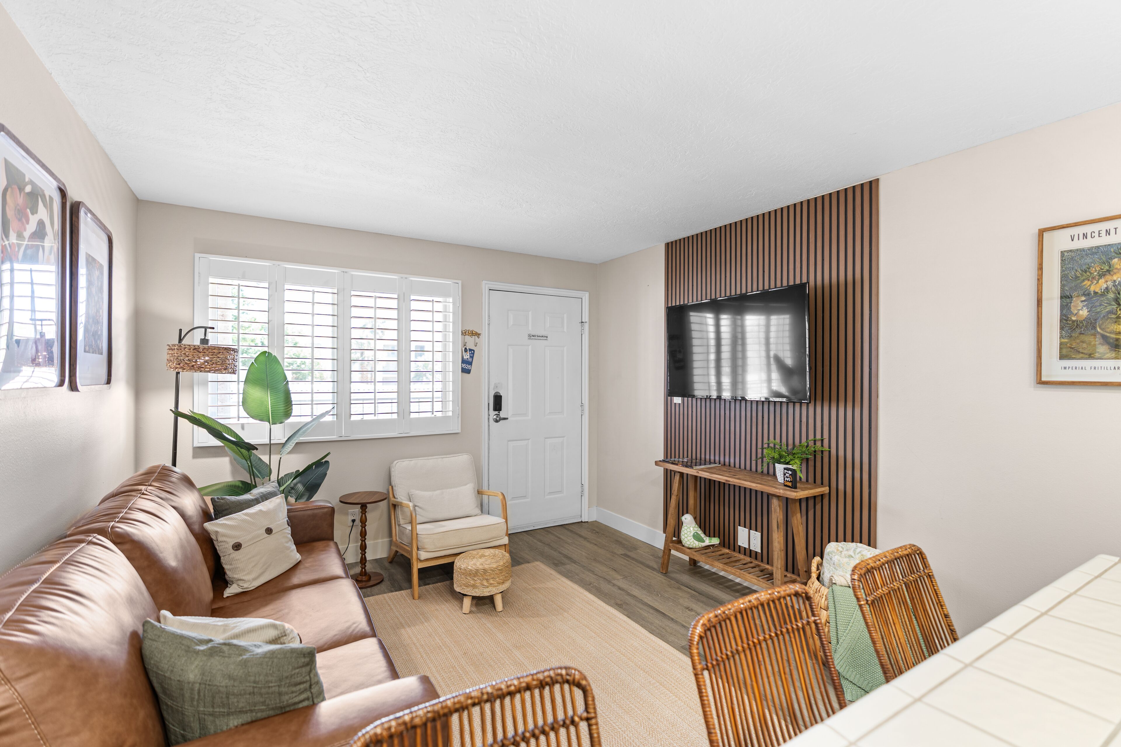 A living room with a brown leather sofa, a television mounted on a wooden wall, a dining table with chairs, and a door with a nameplate.