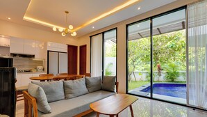 A modern living room with a sofa, wooden table, and large windows offering a view of a swimming pool and greenery. - Villa Ease - Nusa Dua (Nusa Dua)