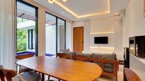 A modern living room with a wooden dining table, comfortable seating, a flat-screen TV, and a large window offering a view of greenery. - Villa Ease - Nusa Dua (Nusa Dua)
