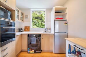 Eine kleine Küche mit Waschmaschine, Kühlschrank und einem Fenster mit Blick auf Grünflächen.