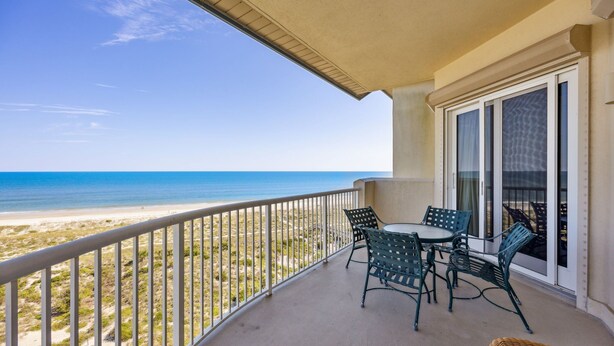 A balcony with a view of the beach, a table with chairs, and a wicker chair. - Experience oceanfront luxury at 32 Ocean Place, a cozy 2-bedroom condo in Fernandina Beach. (Fernandina Beach)