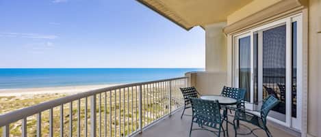 A balcony with a view of the beach, a table with chairs, and a wicker chair.