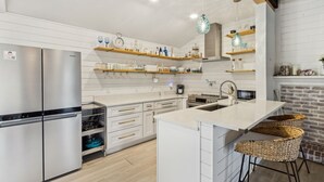 A modern kitchen with a stainless steel refrigerator, white cabinetry, wooden shelves, and a central island with bar stools. - Charming 3BR home in Fernandina Beach with wooded views and space for 8! (Fernandina Beach)