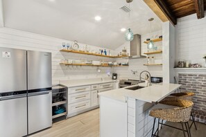 A modern kitchen with a stainless steel refrigerator, white cabinetry, wooden shelves, and a central island with bar stools. - Charming 3BR home in Fernandina Beach with wooded views and space for 8! (Fernandina Beach)