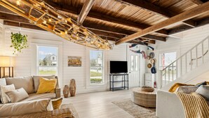 A spacious living room with a high ceiling, wooden beams, a television, wicker furniture, and a staircase. - Charming 3BR home in Fernandina Beach with wooded views and space for 8! (Fernandina Beach)
