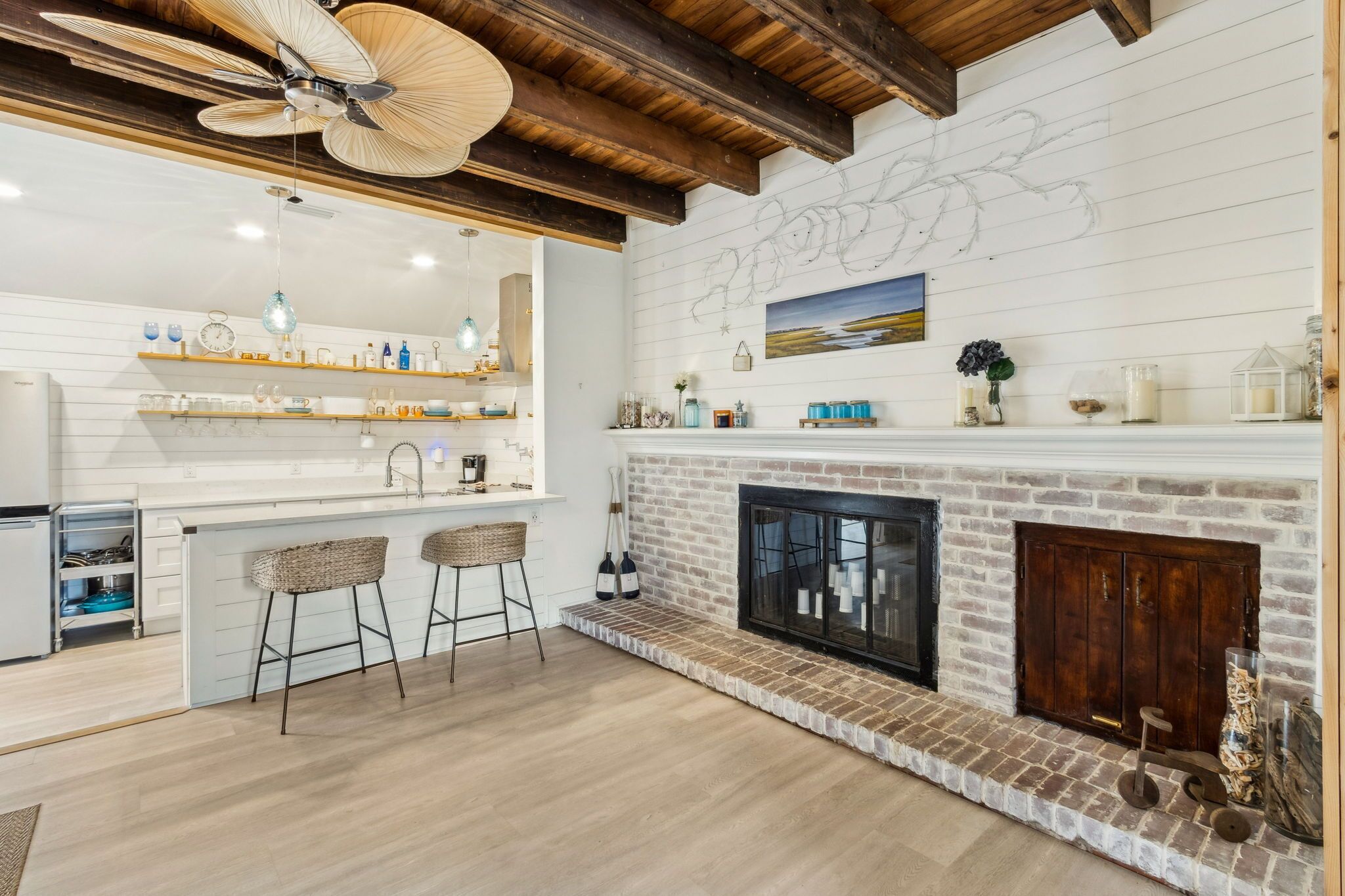 A modern kitchen with a white backsplash, wooden beams, a brick fireplace, and a bar area with stools.