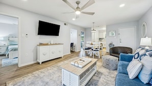 A spacious living room with a ceiling fan, a flat-screen TV mounted on the wall, a dining area with a table and chairs, and a cozy seating area with a blue sofa and patterned cushions.