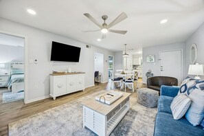 Ein geräumiges Wohnzimmer mit Deckenventilator, einem an der Wand befestigten Flachbildfernseher, einem Essbereich mit Tisch und Stühlen sowie einer gemütlichen Sitzecke mit blauem Sofa und gemusterten Kissen.