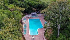 An aerial view of a rectangular swimming pool surrounded by lounge chairs and a small building, likely a changing room or storage, amidst dense greenery.