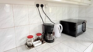 Una cocina con paredes de azulejos blancos, una cafetera negra, un hervidor blanco y un microondas negro. Hay dos frascos con tapas rojas sobre la encimera.