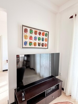 Un salon doté d’un grand téléviseur à écran plat, d’une collection de masques colorés encadrés accrochés au mur et d’un meuble de rangement en bois foncé placé en dessous.