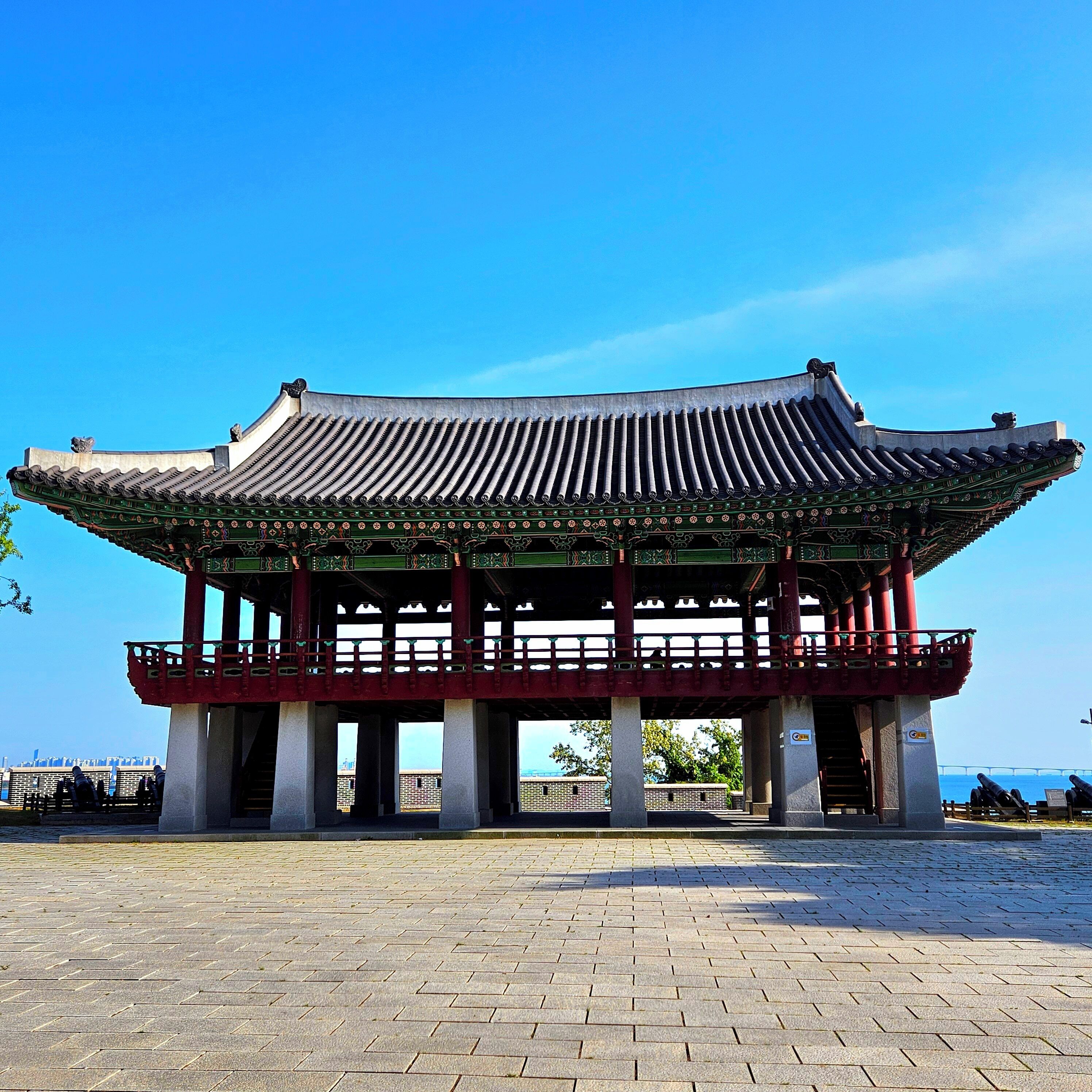 A traditional Korean pavilion with a tiled roof and a balcony, situated on a paved area with a clear view of the sea in the background.
