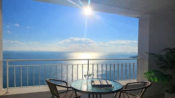 A balcony with a glass table and two chairs, overlooking a body of water and a distant bridge.