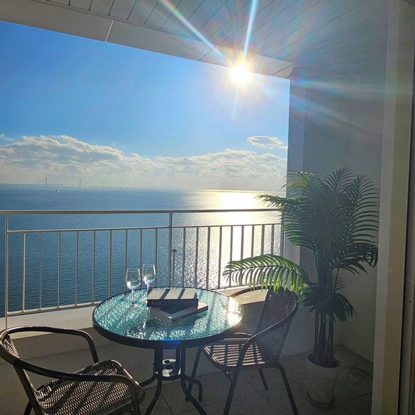 A balcony with a glass table, two chairs, a potted plant, and a view of the sea and a bridge. - SainTree Hotel (Incheon)
