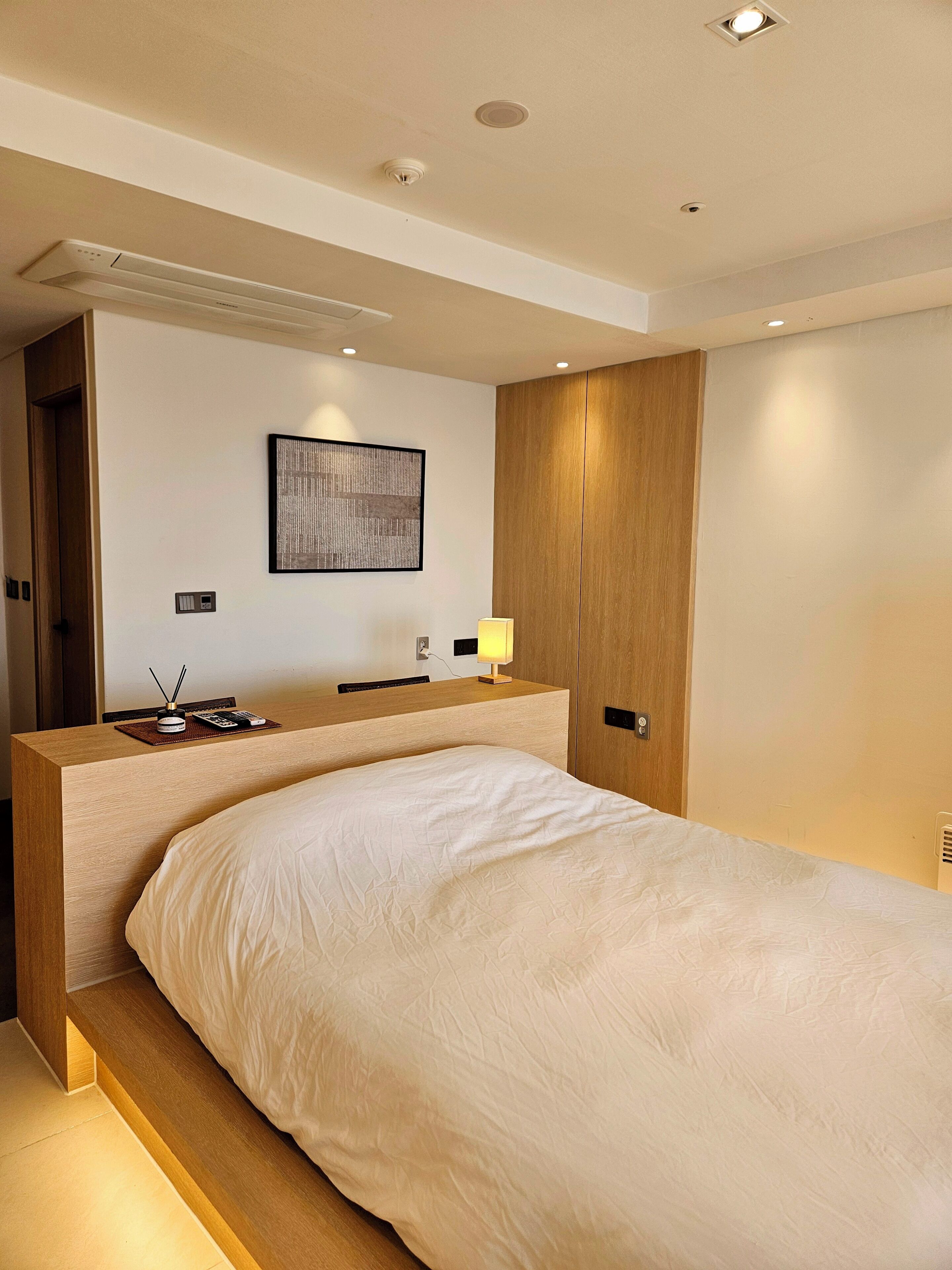 Une chambre d’hôtel moderne avec un grand lit, un bureau en bois, une lampe fixée au mur et une œuvre d’art encadrée.