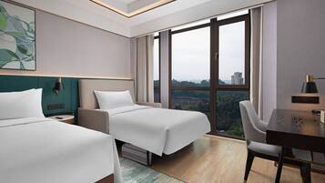 A hotel room with two beds, a desk, and a chair. There is a large window with a view of buildings and trees.