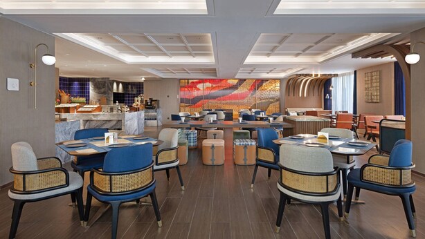 A modern restaurant with a central dining area featuring round tables and blue chairs, a bar area with stools, and a large abstract mural on the back wall. - Hilton Garden Inn Leshan Shizhong (Leshan)