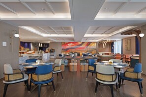 A modern restaurant with a central dining area featuring round tables and blue chairs, a bar area with stools, and a large abstract mural on the back wall. - Hilton Garden Inn Leshan Shizhong (Leshan)