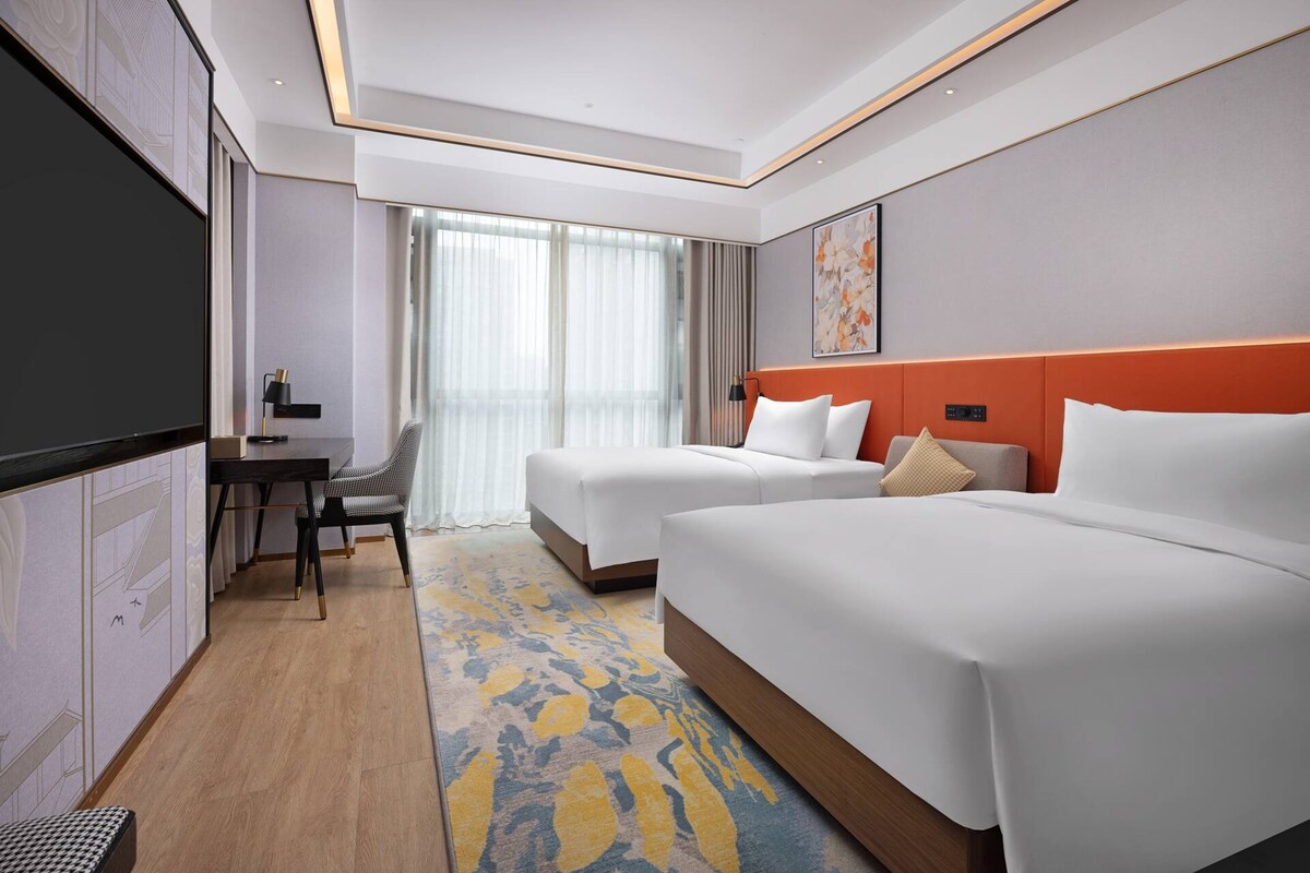 A hotel room with two beds, a desk, a chair, a TV, and a large window with curtains.