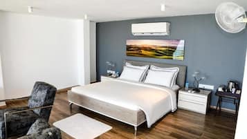 A modern bedroom with a large bed, a grey upholstered chair, a bedside table with a lamp, and a wall-mounted air conditioner.