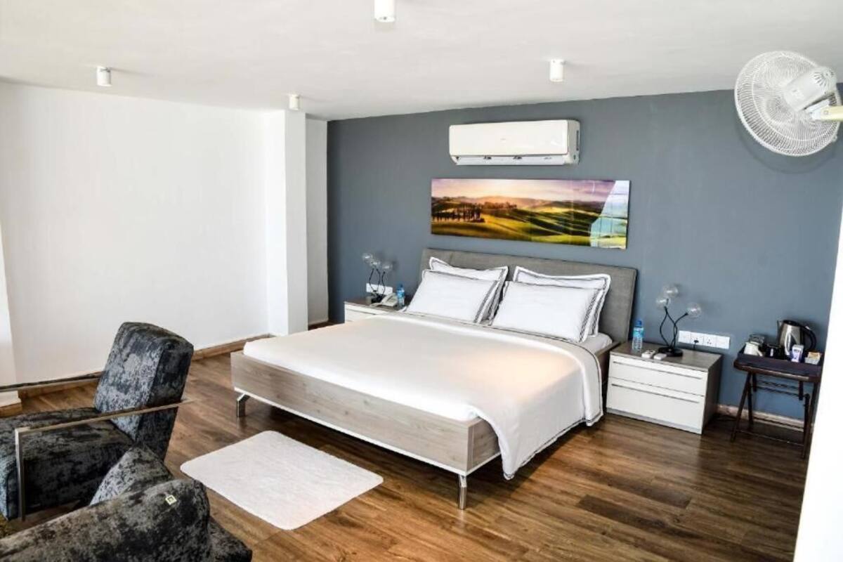 A modern bedroom with a large bed, a grey upholstered chair, a bedside table with a lamp, and a wall-mounted air conditioner.