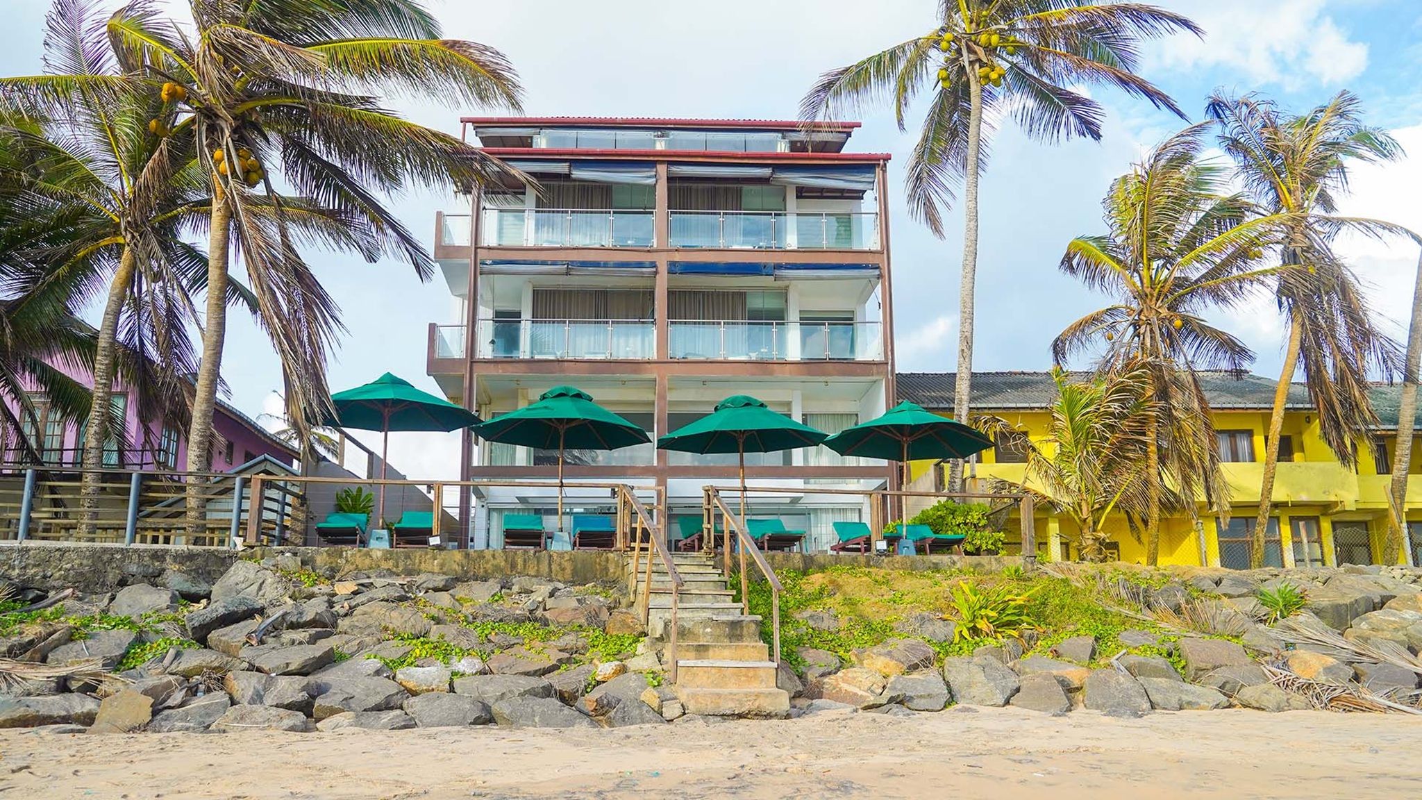 A multi-story hotel with balconies, outdoor seating with umbrellas, and a rocky foreground.