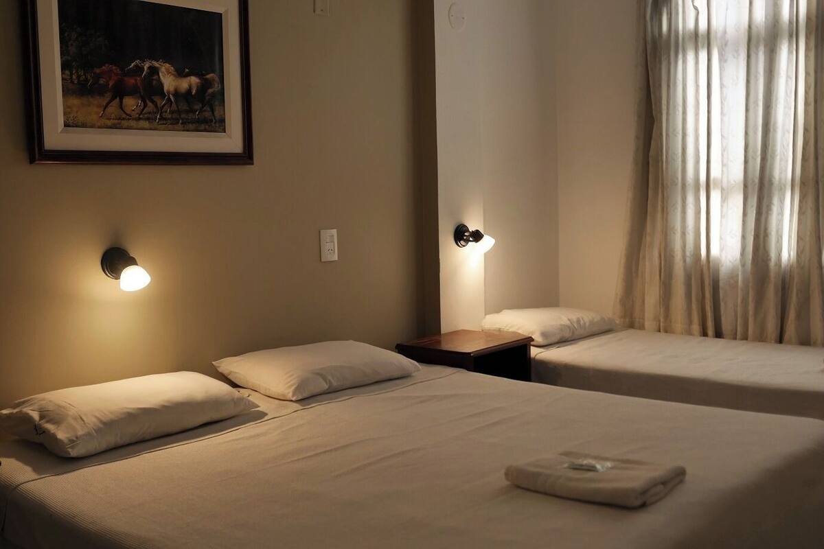 A hotel room with two beds, each with a pillow and a bedside table. There is a painting on the wall above the beds.