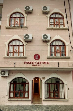 La façade d’un hôtel présente de nombreuses fenêtres en arc de cercle, un logo central et le nom « Paseo Cuemés Hotel » affiché en évidence.
