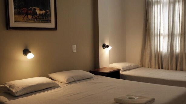 A hotel room with two beds, each with a pillow and a bedside table. There is a painting on the wall above the beds. - Paseo Guemes Hotel (Salta)