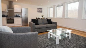 A modern living room with a grey sofa, a glass coffee table, and a kitchen area with a fridge, microwave, and stove. - Celador Apartments - Sussex House (Reading)