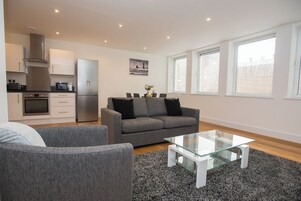 A modern living room with a grey sofa, a glass coffee table, and a kitchen area with a fridge, microwave, and stove.