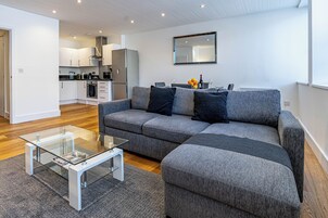A modern living room with a grey sectional sofa, a glass coffee table, and a kitchen in the background.