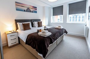 A neatly arranged hotel room with a large bed, a bedside table, a nightstand, and a window with a view of buildings.