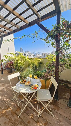 Un'area pranzo all'aperto coperta, con un tavolo apparecchiato per due, circondata da piante in vaso e con vista sulla città.