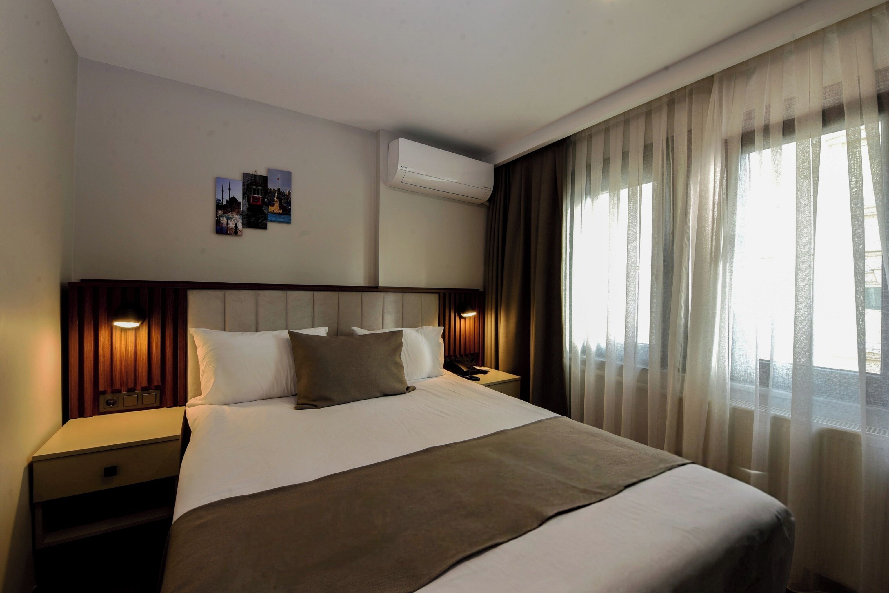 A hotel room with a large bed, a bedside table, an air conditioning unit, and a window with sheer curtains.
