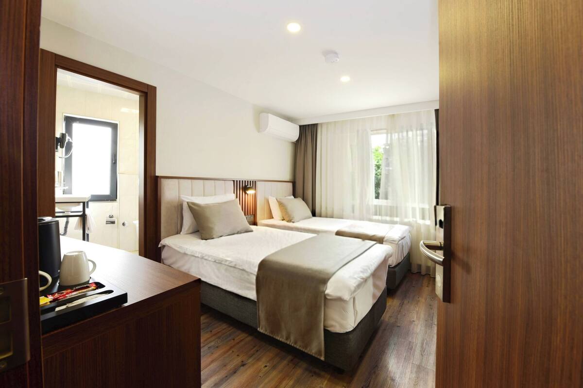 Ein Hotelzimmer mit zwei Betten, einem Holztisch, einer Kaffeemaschine und einem Fenster mit Vorhängen.