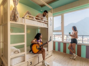 A dormitory room with bunk beds, a window with a view, and a person playing a guitar. - Moustache Rishikesh Luxuria (Narendranagar)