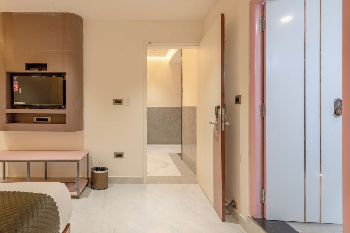 A hotel room with a flat-screen TV mounted on a wall, a bench, a trash can, and a wardrobe with a mirror.