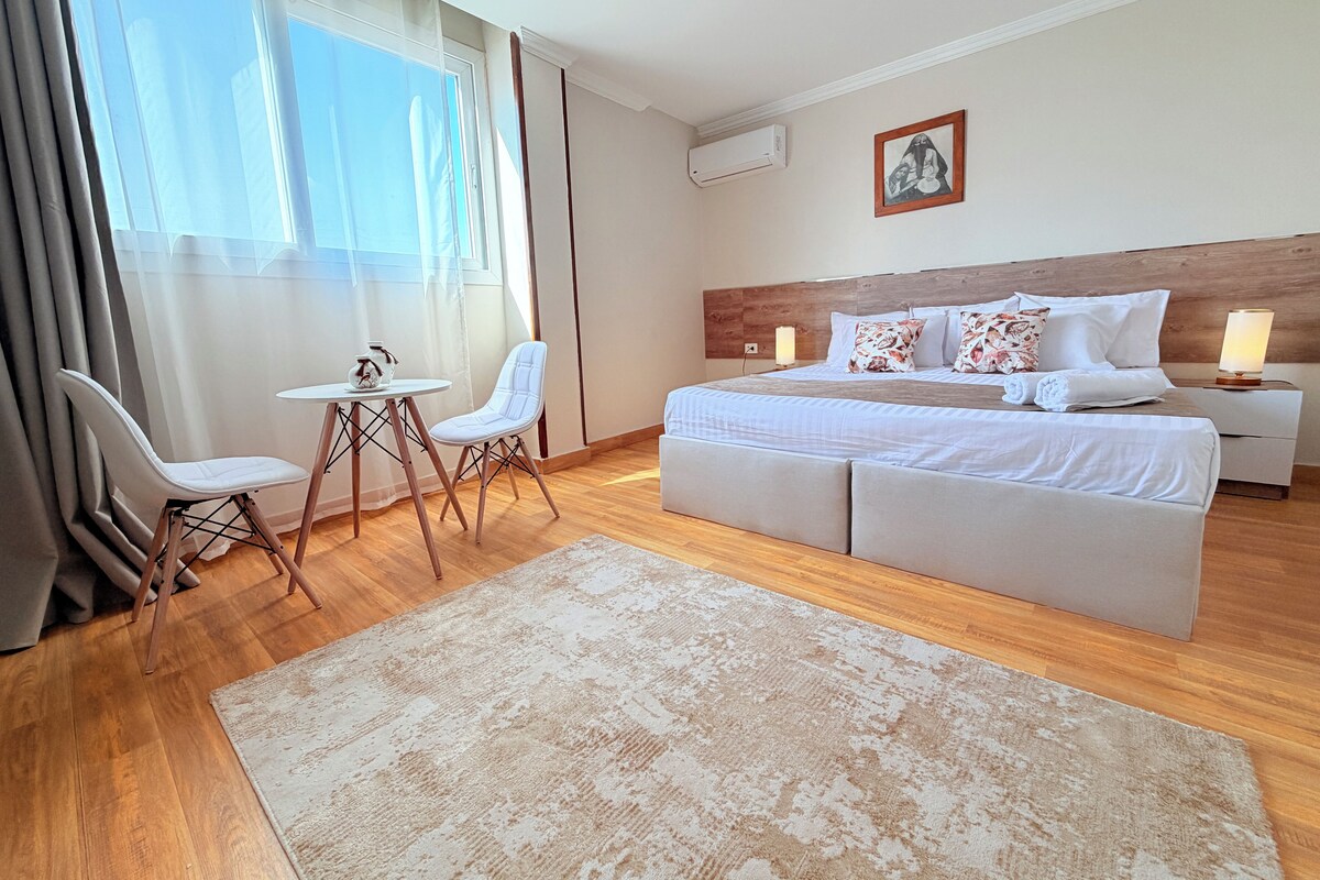 A modern hotel room with a large bed, a small table, two chairs, a large window with curtains, and a wooden floor.