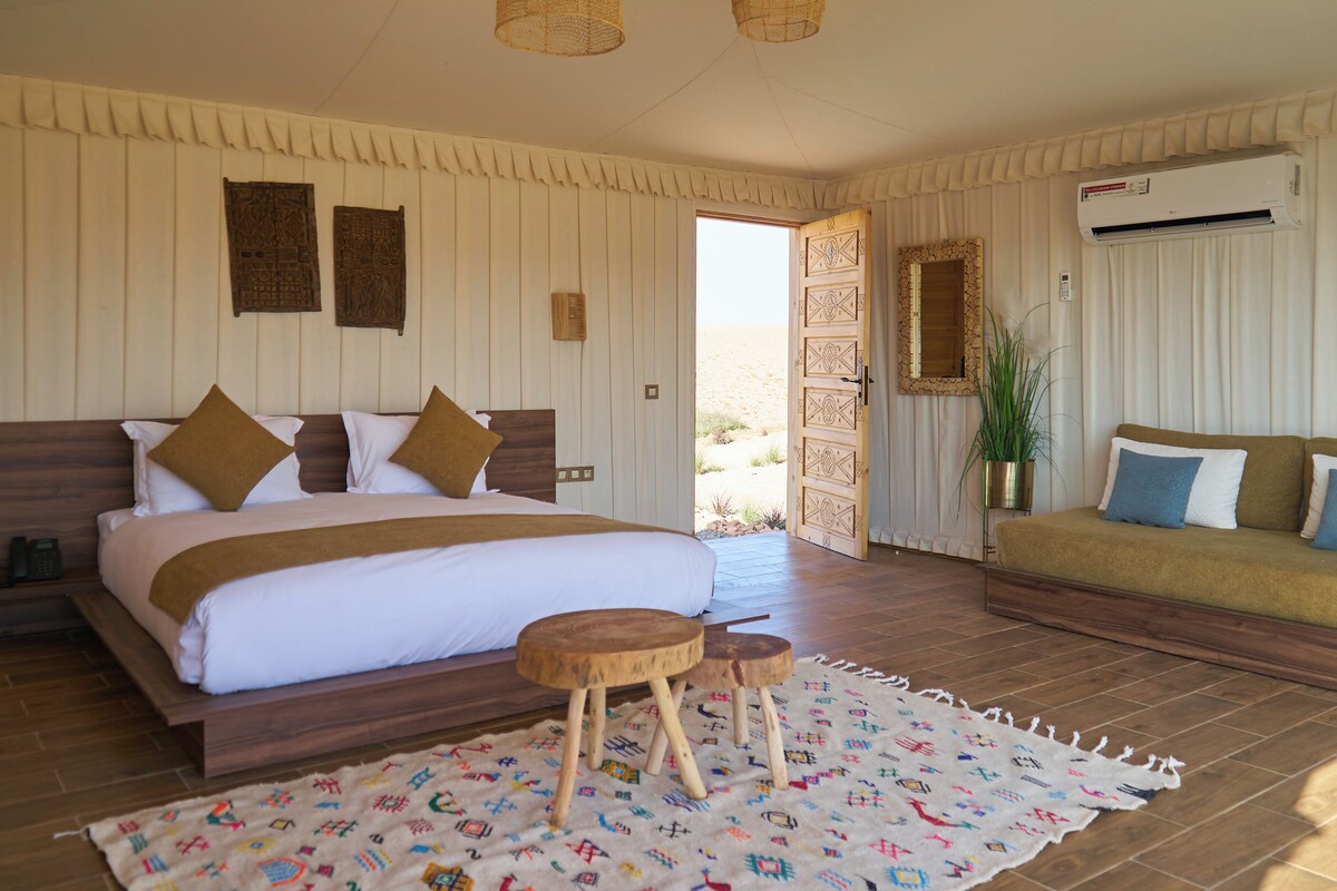 A bedroom with a large bed, a wooden bench, a patterned rug, and a view of the desert through an open door.