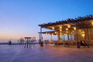 An outdoor dining area with tables and chairs, a pergola with hanging lights, and a view of the sea. - Stellar Desert Camp (Marrakech)