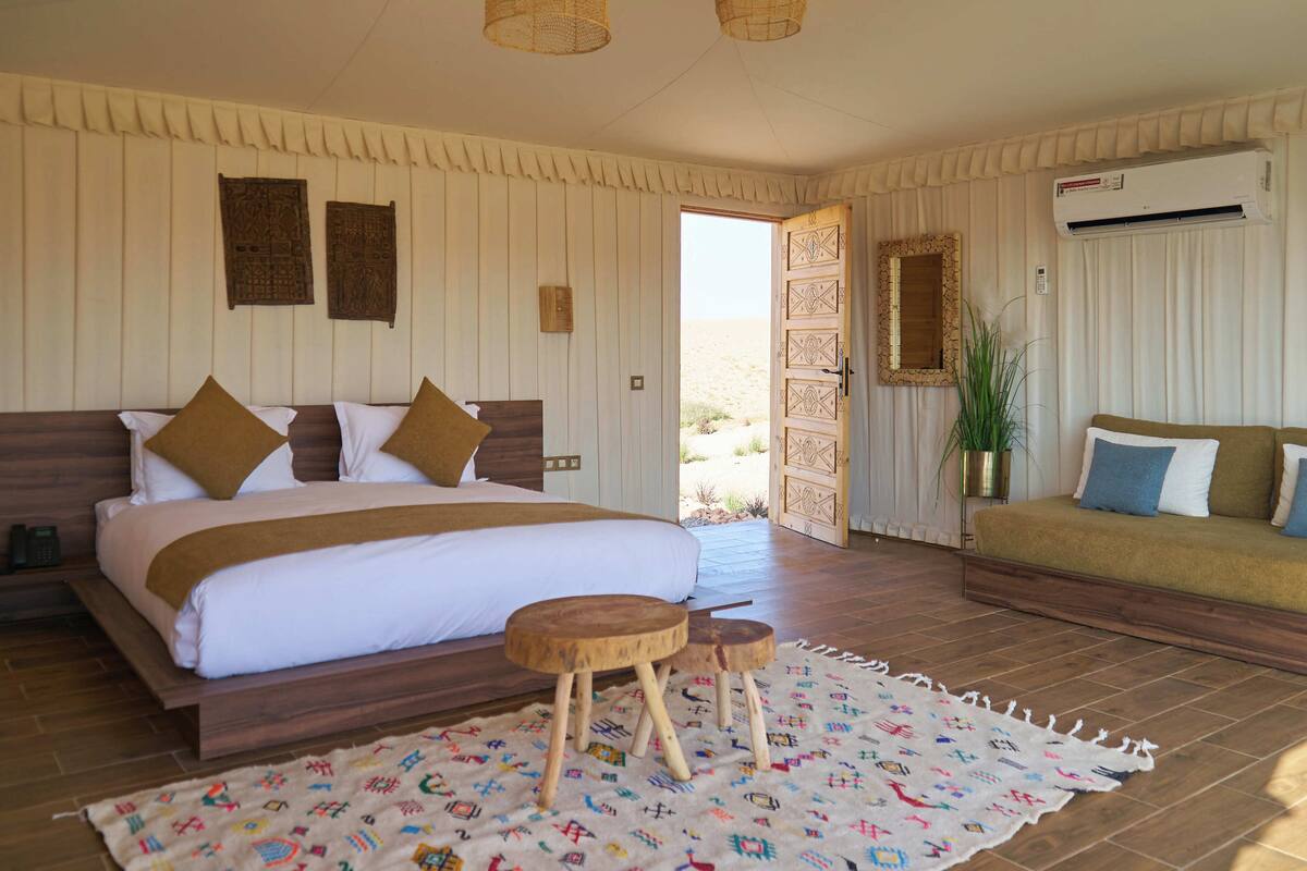 A bedroom with a large bed, a wooden bench, a patterned rug, and a view of the desert through an open door.