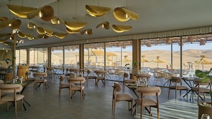 A spacious dining area with multiple tables and chairs, large windows offering a view of the desert, and hanging gold-colored pendant lights. - Stellar Desert Camp (Marrakech)
