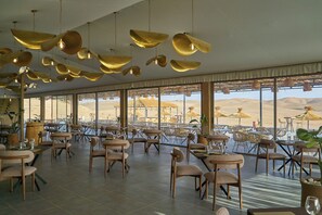 Un comedor amplio con varias mesas y sillas, ventanas grandes que ofrecen vistas al desierto, y lámparas colgantes doradas.