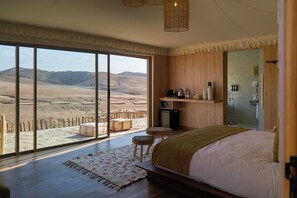 A bedroom with a large sliding glass door leading to a desert view, a bed with a brown throw, a small stool, and a nightstand with toiletries. - Stellar Desert Camp (Marrakech)