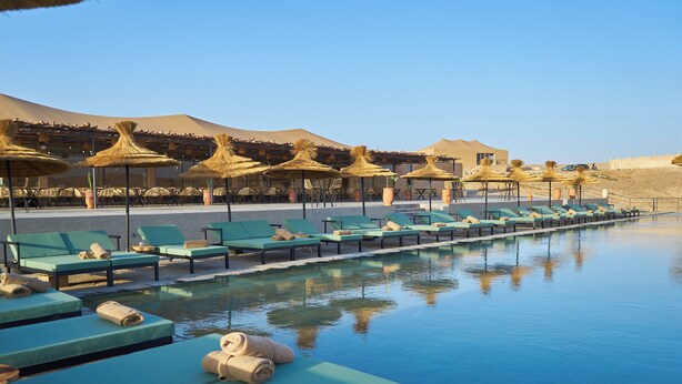 A poolside area with lounge chairs and thatched umbrellas, a clear sky, and a desert landscape in the background. - Stellar Desert Camp (Marrakech)