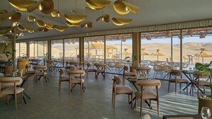 A spacious dining area with multiple tables and chairs, large windows offering a view of the desert, and hanging gold-colored pendant lights. - Stellar Desert Camp (Marrakech)