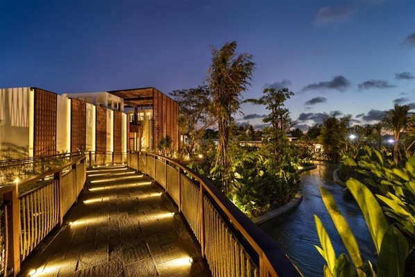 Vue nocturne d'un hôtel moderne doté d'une façade en verre, d'une terrasse en bois et d'un élément décoratif aquatique.